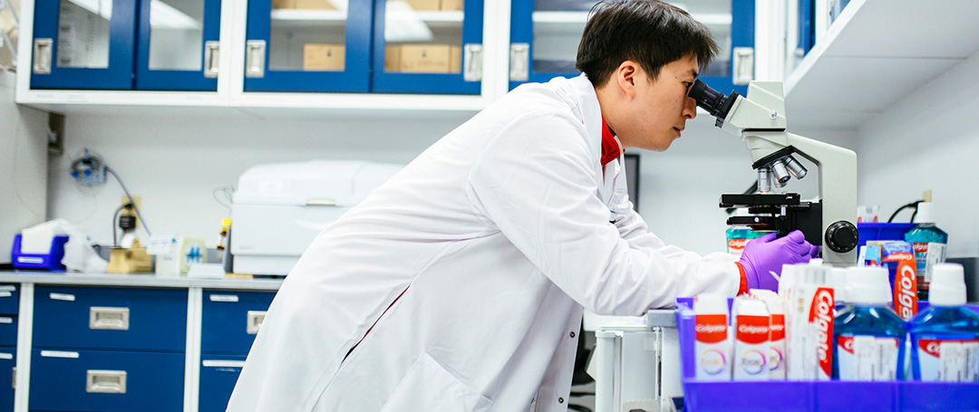 Un hombre mira por un microscopio en un laboratorio junto a un grupo de productos Colgate.
